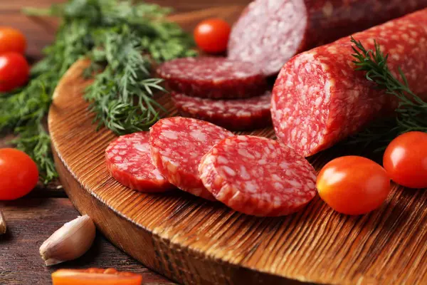Farklı tütsülenmiş sosisler, ahşap masada kiraz domatesleri ve baharatlar, yakın plan.