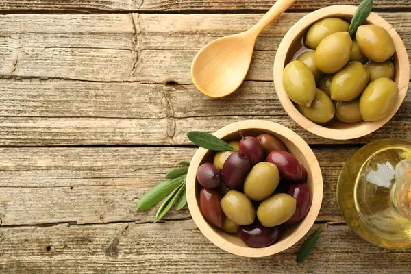 Lezzetli zeytinler kaselerde, yeşil yapraklar, kaşık ve yağ ahşap masada, düz yerde. Metin için boşluk