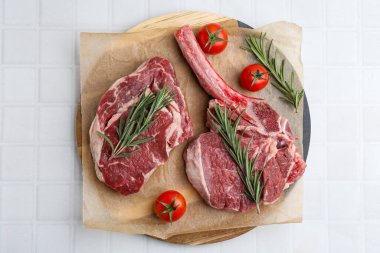 Pieces of raw beef, rosemary and tomatoes on white tiled table, top view