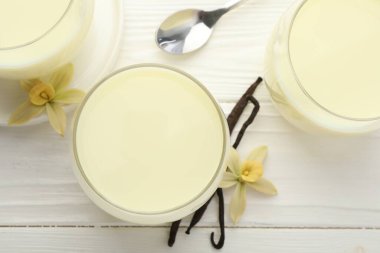 Bardaklarda vanilyalı süt, kapsüller ve çiçekler beyaz ahşap masada, düz yerde