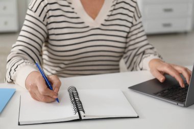 İnternette bilgisayar kullanarak öğrenen ve içerideki beyaz masada notlar alan bir kadın. Kendi kendine çalışma.