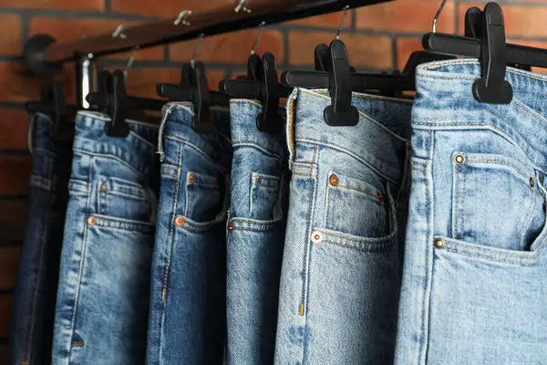 Many stylish jeans hanging from rack near brick wall, closeup
