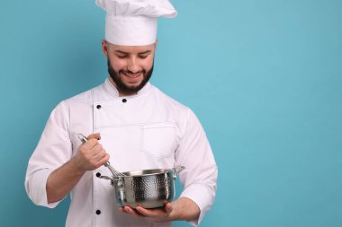 Üniformalı mutlu pastacı elinde çırpıcı ve açık mavi arka planda pişirme kabı tutuyor.