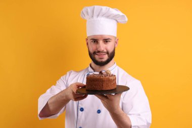 Üniformalı mutlu pastacı turuncu arka planda çikolatalı pasta tutuyor.