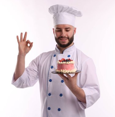 Üniformalı mutlu pastacı elinde böğürtlenli lezzetli bir pasta tutuyor ve beyaz arka planda güzel bir jest yapıyor.