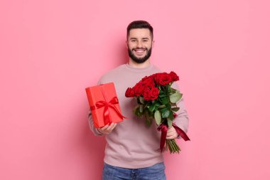 Kırmızı gül buketi ve pembe arka planda hediye kutusu olan mutlu bir adam.