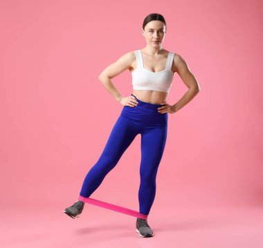 Spor kıyafetli bir kadın pembe arka planda elastik bantla egzersiz yapıyor.