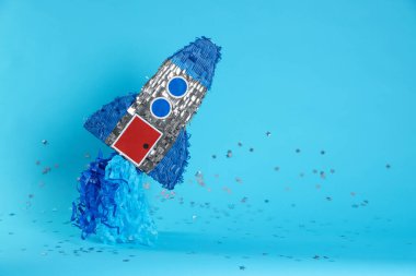 Açık mavi arkaplanda roket şekilli pinata ve konfeti, metin için boşluk