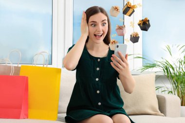 İkramiye. Evde cep telefonu kullanan sürpriz bir kadın. Hediye kutuları ve yüzde işaretleri cihazdan uçuyor