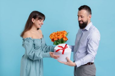 Bir erkek, açık mavi arka planda sevgili karısına güzel laleler ve hediye kutusu sunar.
