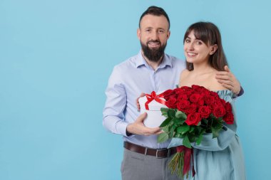 Kırmızı gülleri ve açık mavi arka planda hediye kutusu olan sevimli bir çift.