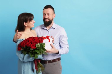 Kırmızı gülleri ve açık mavi arka planda hediye kutusu olan sevimli bir çift.