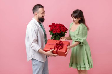 Kırmızı gül buketini ve hediye kutusunu pembe arka plandaki sevgilisine sunan bir adam.