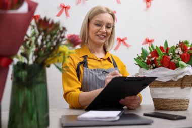 Elinde pano olan bir işyeri sahibi çiçekçi dükkanındaki masada notlar alıyor.