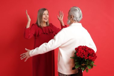 Kırmızı arka planda, mutlu karısı için bir buket gül saklayan adam.