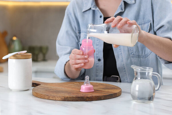 Mother making baby formula in feeding bottle at table indoors, closeup