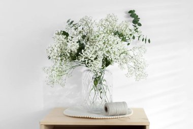 Bouquet of beautiful gypsophila flowers and eucalyptus branches in vase on chest of drawers near white wall