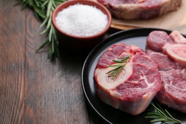 Pieces of raw beef, rosemary and salt on wooden table, closeup. Space for text