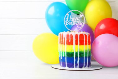 Lezzetli gökkuşağı doğum günü pastası. Masada beyaz ahşap arka planda balonlar, metin için yer var.