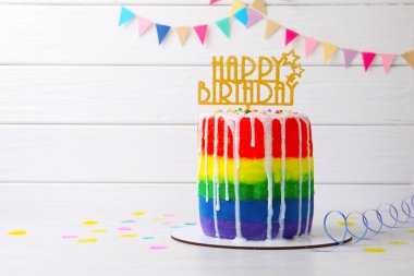 Lezzetli gökkuşağı doğum günü pastası. Masada beyaz ahşap arka planda konfeti ve yazı için yer var.