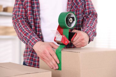 Karton kutuyu kapalı bir yere kapatmak için yapıştırıcılı bant kullanan bir adam.