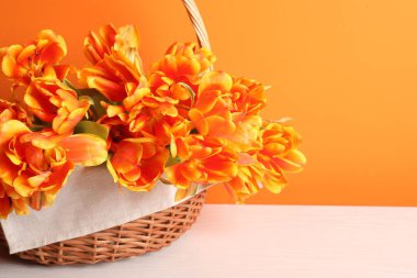 Hasır sepetteki güzel laleler beyaz ahşap masada turuncu arka planda, yakın plan. Metin için boşluk