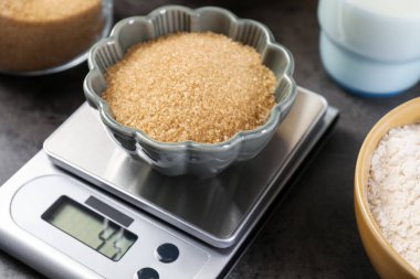 Dijital mutfak terazisi, kahverengi şeker kasesi ve gri masadaki diğer ürünler, yakın plan.