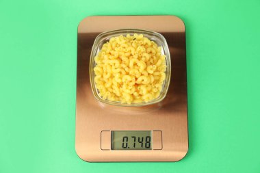 Electronic kitchen scale with bowl of raw pasta on green background, top view