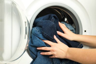 Kadın çamaşır makinesine kot pantolon koyuyor, yakın plan.