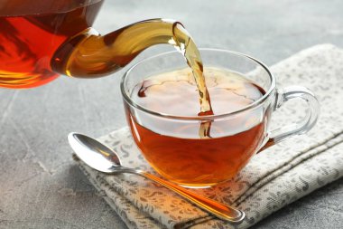 Gri masada çaydanlıktan fincana taze demlenmiş çay dökülüyor, yakın plan.