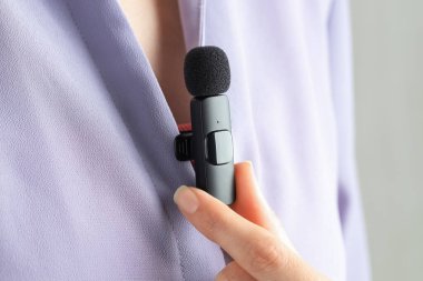 Kadın açık gri arka planda gömleğine mikrofon takıyor, yakın plan.