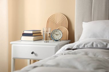 Yatağın yanında kitap, dekor, bir bardak su ve çalar saat olan komodin.
