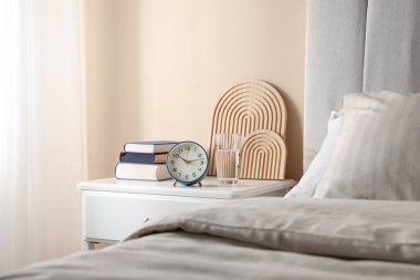 Yatağın yanında kitap, dekor, bir bardak su ve çalar saat olan komodin.