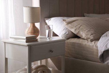 Yatağın yanında kitap, lamba ve bir bardak su olan komodin.