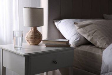 Yatağın yanında kitap, lamba ve bir bardak su olan komodin.