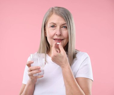 Bir bardak suyla mutlu bir kadın pembe arka planda kolajen hapı alıyor.