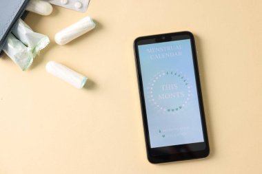 Dönem takvimi uygulamalı akıllı telefon ve bej arka planda adet ürünleri, düz yatış