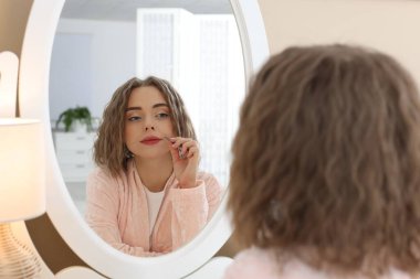 Güzel bir kadın evde aynanın yanında cımbızla bıyığını koparıyor.