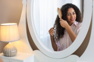 Gülümseyen genç bir kadın evde aynanın yanında kıvırcık saçlarını tarıyor.