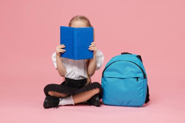 Sırt çantalı liseli kız pembe arka planda kitap okuyor.