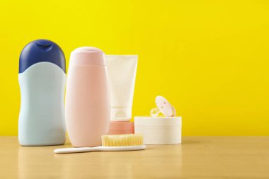 Bebek kozmetik ürünleri ve banyo aksesuarları ahşap masada, sarı arka planda. Metin için boşluk