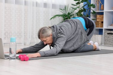 Evde dizüstü bilgisayar başında egzersiz yapan yaşlı bir kadın.