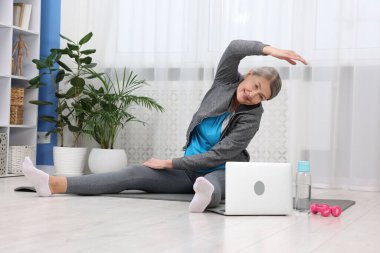 Evde laptopun yanında egzersiz yapan gülümseyen yaşlı kadın.