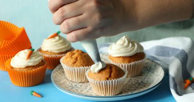 Mavi masada nefis havuçlu keklerin üzerine krem şanti sıkan bir kadın.