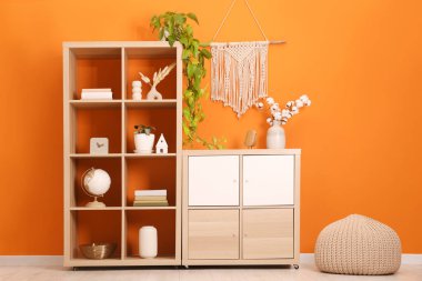 Wooden shelving unit with different home decor, houseplant and pouf near orange wall indoors
