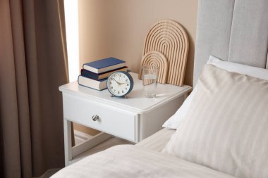 Yatağın yanında kitap, dekor, bir bardak su ve çalar saat olan komodin.