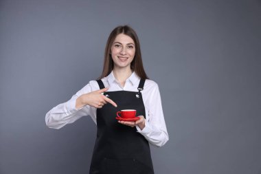 Gülümseyen baristanın portresi gri arka planda bir fincan sıcak içeceğe işaret ediyor.