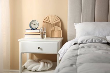 Yatağın yanında kitap, dekor, bir bardak su ve çalar saat olan komodin.