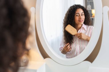 Gülümseyen genç bir kadın evde aynanın yanında kıvırcık saçlarını tarıyor, seçici bir şekilde odaklanıyor.