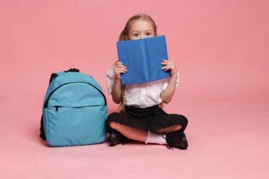 Sırt çantalı liseli kız pembe arka planda kitap okuyor.